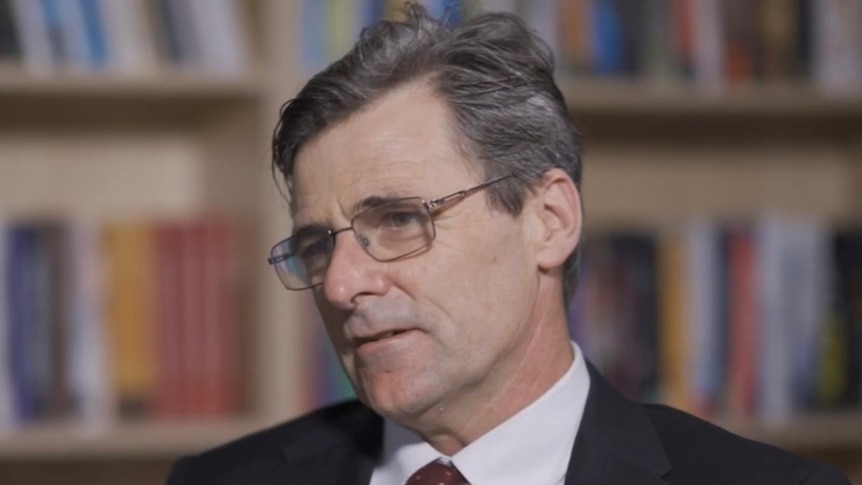 A middle-aged man with grey hair wearing glasses and a suit in an interview with blurred book shelf in background