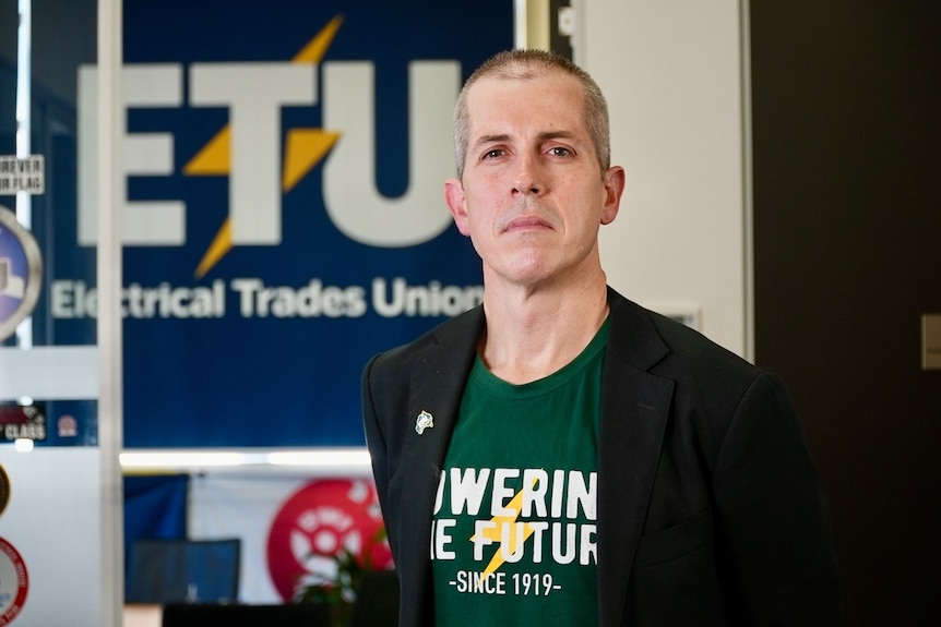 Michael Wright wearing a dark green shirt that says "Powering the Future", standing in front of an Electrical Trades Union sign.