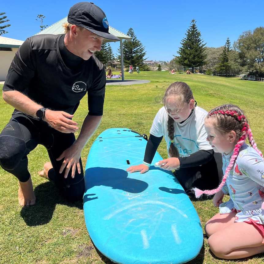 Harley down on one knee helping two young girls wax a surfboard.