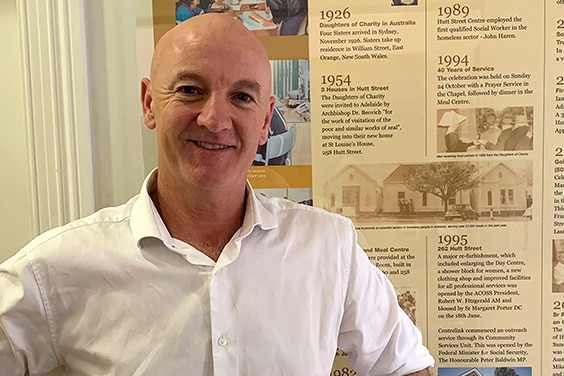 A man smiles at the camera in front of a wall.