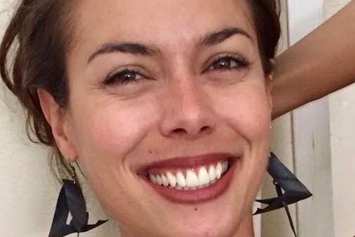 A woman with brown hair, triangular earrings and a blue dress holds a hand behind her head while she smiles widely.