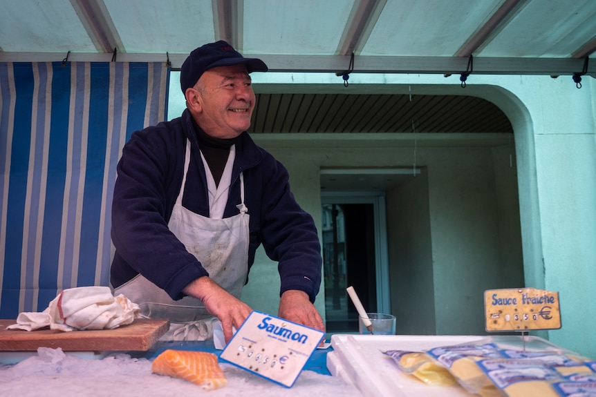 A fishmonger in an apron smiles at a customer who is out of shot.