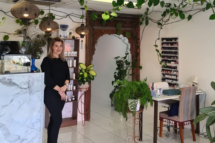 Young woman with blonde hair smiles as she stands by a marble counter in a small beauty salon adorned with plenty of plants.