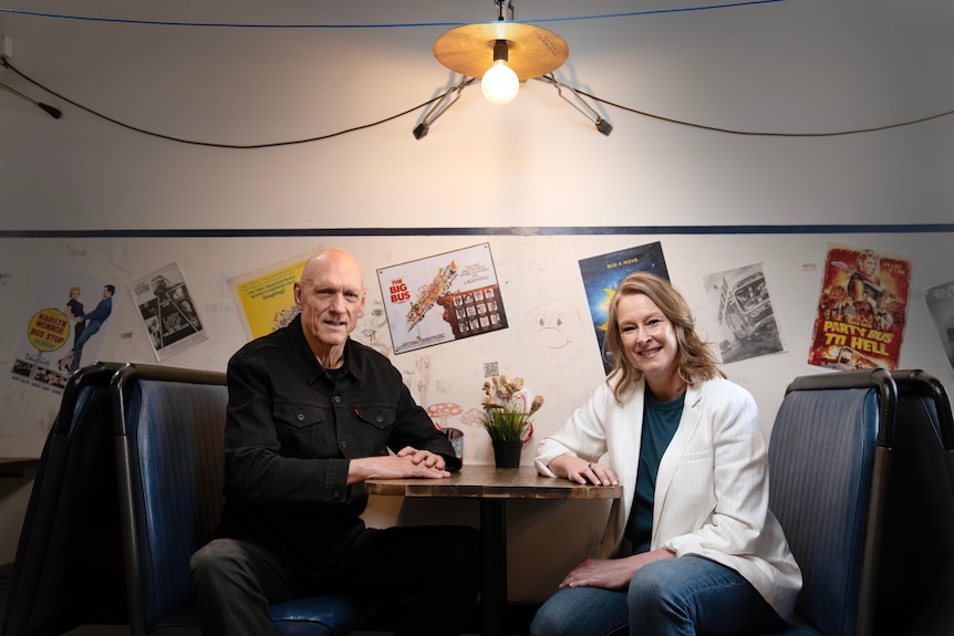 Peter Garrett and Leigh Sales sitting at a booth table, posters behind
