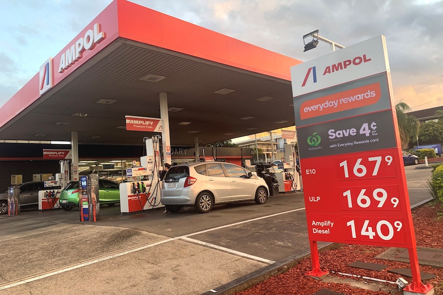 Cars in the driveway of an Ampol petrol station with a sign showing the price per litre for fuel
