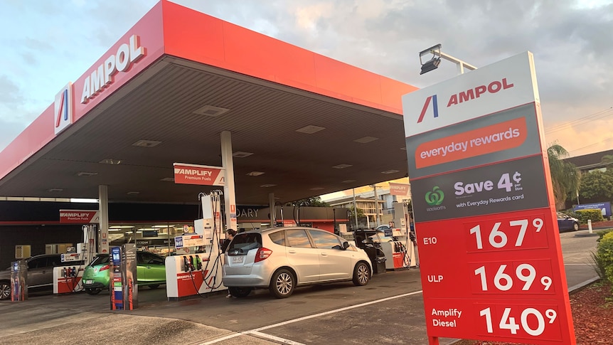 Cars in the driveway of an Ampol petrol station with a sign showing the price per litre for fuel