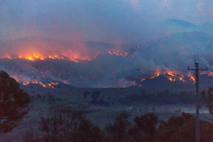 Flames with heavy smoke spread across hilly terrain.