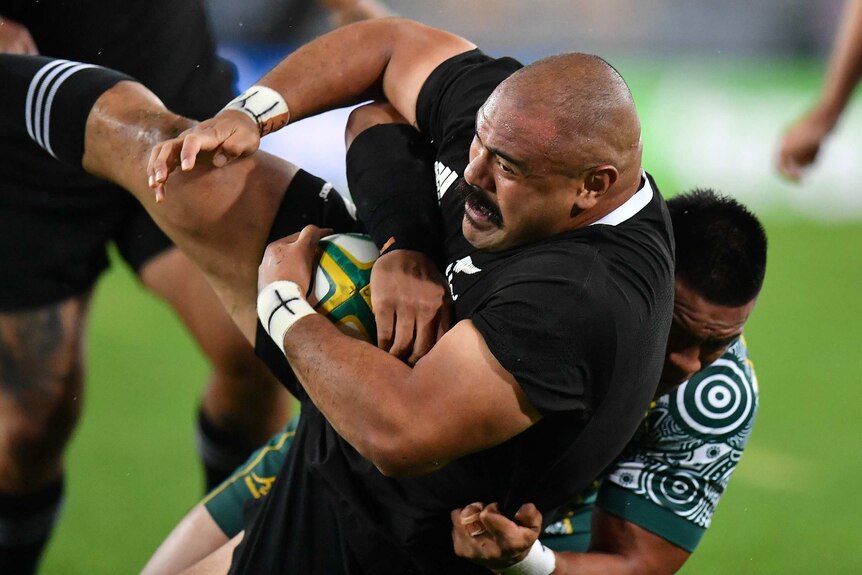 A New Zealand All Blacks player is tackled by a Wallabies opponent as he holds the ball with his right arm.