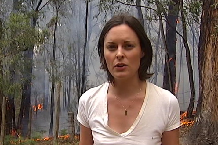 Journalist looking at camera with small flames burning in bush and smoke in background.