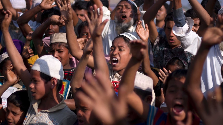 A group of Rohingya children and refugees stand together and raise their arms in the air and shout.