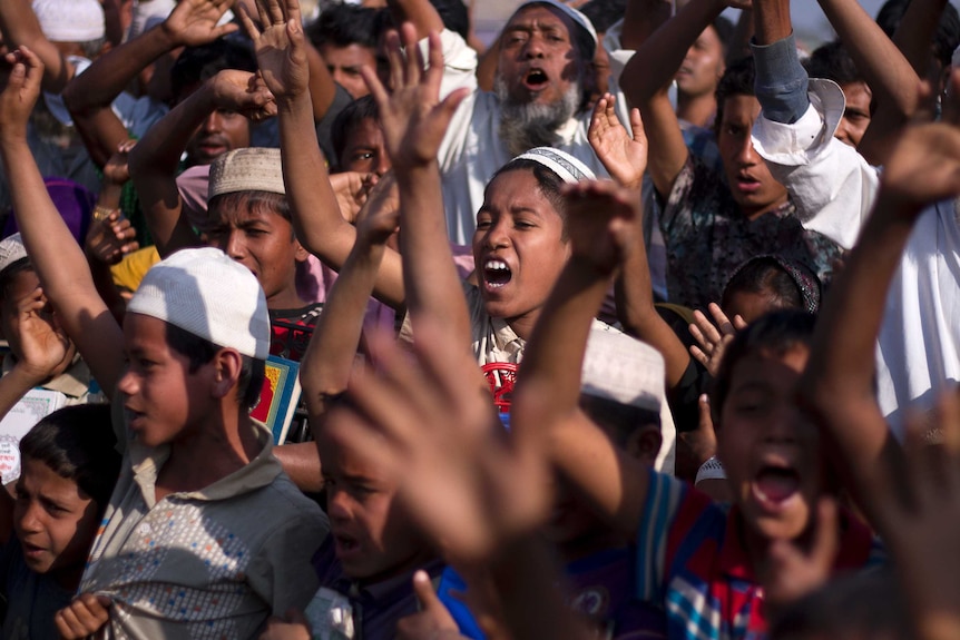 A group of Rohingya children and refugees stand together and raise their arms in the air and shout.