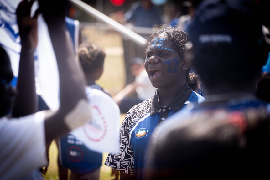 A young woman with blue face paint shouting out across a football field