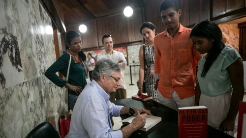 A man sitting at a table signs copies of his book as fans watch on.