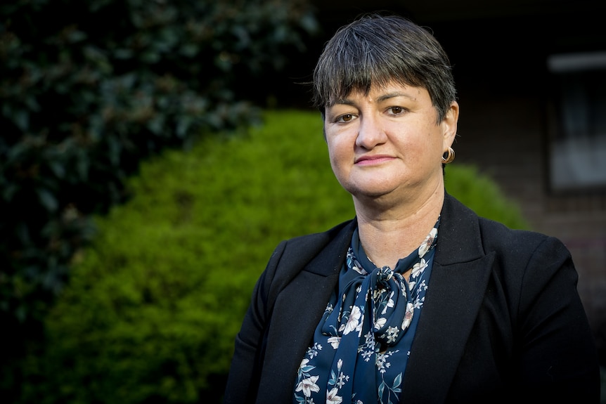 A middle-aged woman with dark grey hair and a fringe, standing outside.