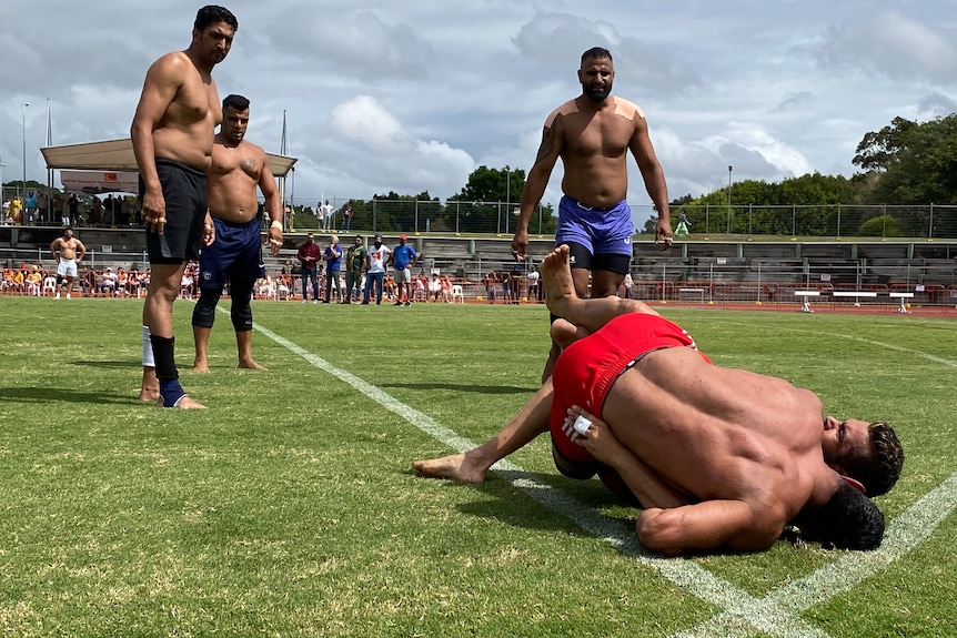 Two shirtless men wrestle on the ground while three other men watch on during an Indian game called Kabbadi.