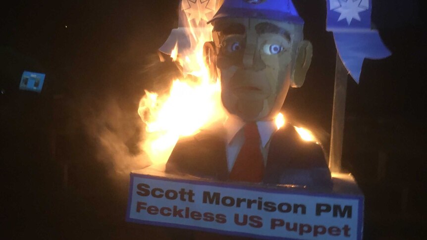 A cardboard effigy of PM Scott Morrison is on fire, two signs read "#freeAssange" and Scott Morrison PM Feckless US puppet