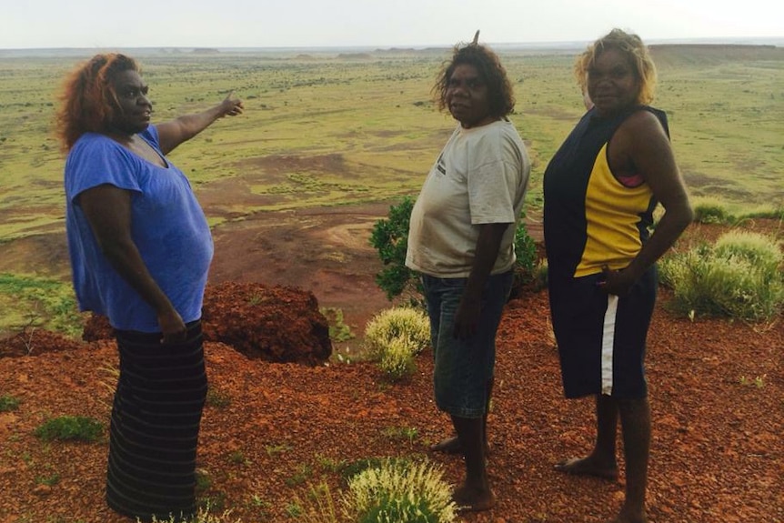 Three Aboriginal women stand in from of a grassy plain at Balgo Hills in the Kimberley, looking at the camera.