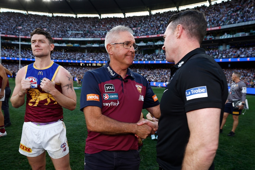 Two AFL coaches shake hands on the MCG after the grand final, while a losing player standing on the left of screen looks away.