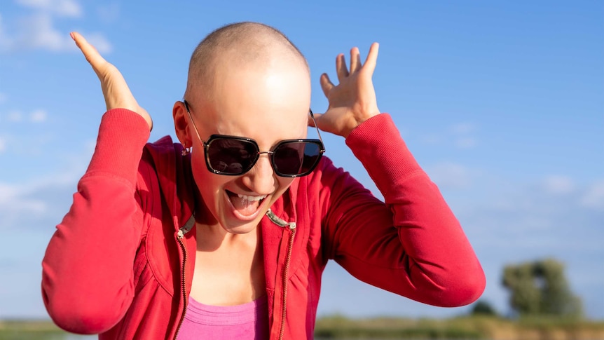 Young woman with a bald head wearing sunglasses and a red jacket with her hands to her ears and mouth open