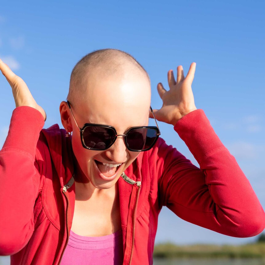 Young woman with a bald head wearing sunglasses and a red jacket with her hands to her ears and mouth open