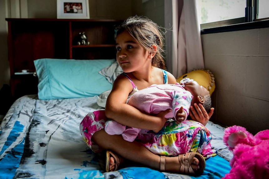 Karla De Lautour nurses a doll while sitting on her bed.