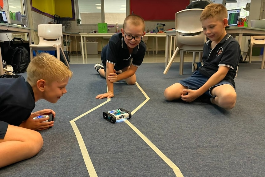 Three young boys watch small robots follow a line in a classroom.