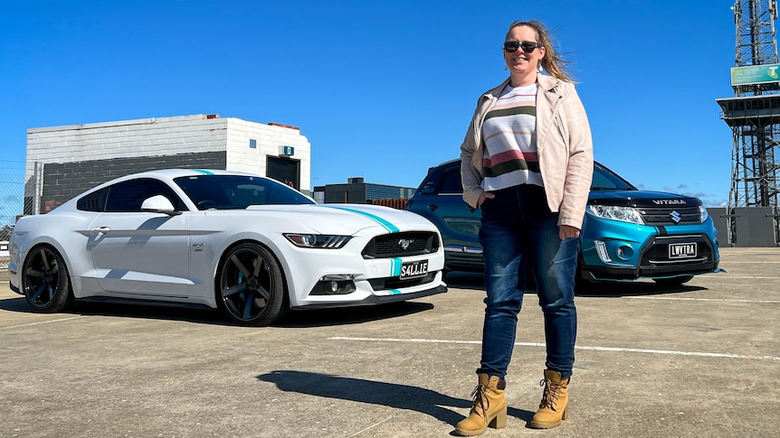 A woman stands in front of her two cars, a white mustang and a teal vitara