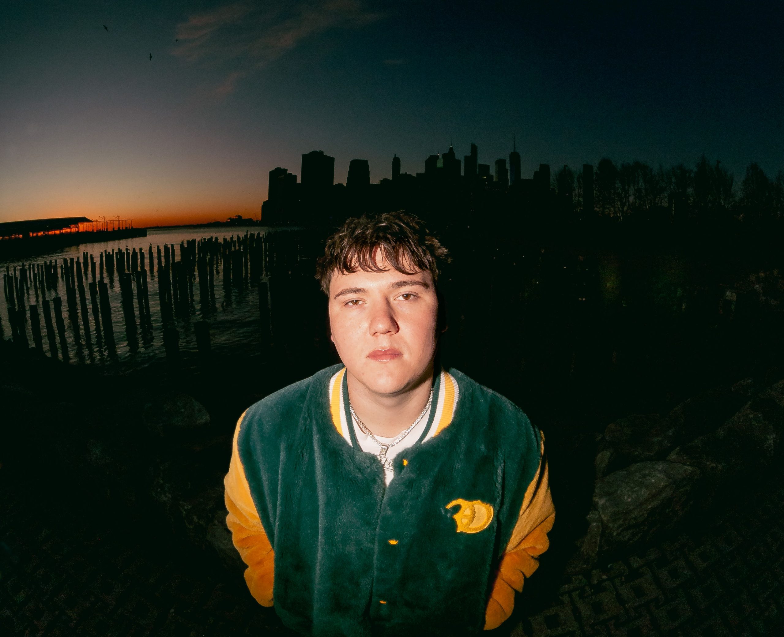 YNG Martyr looks into the camera, with a city at dusk in the background. 