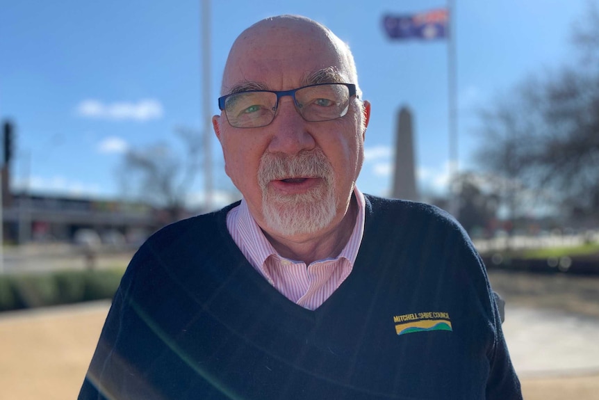 A bald man with a white goatee and glasses standing outside in front of a flag