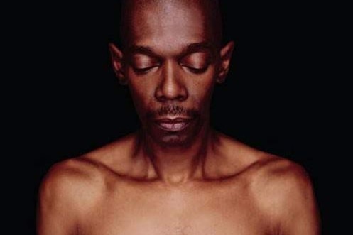 Head and shoulders shot of a bald, topless black man withe eyes downcast