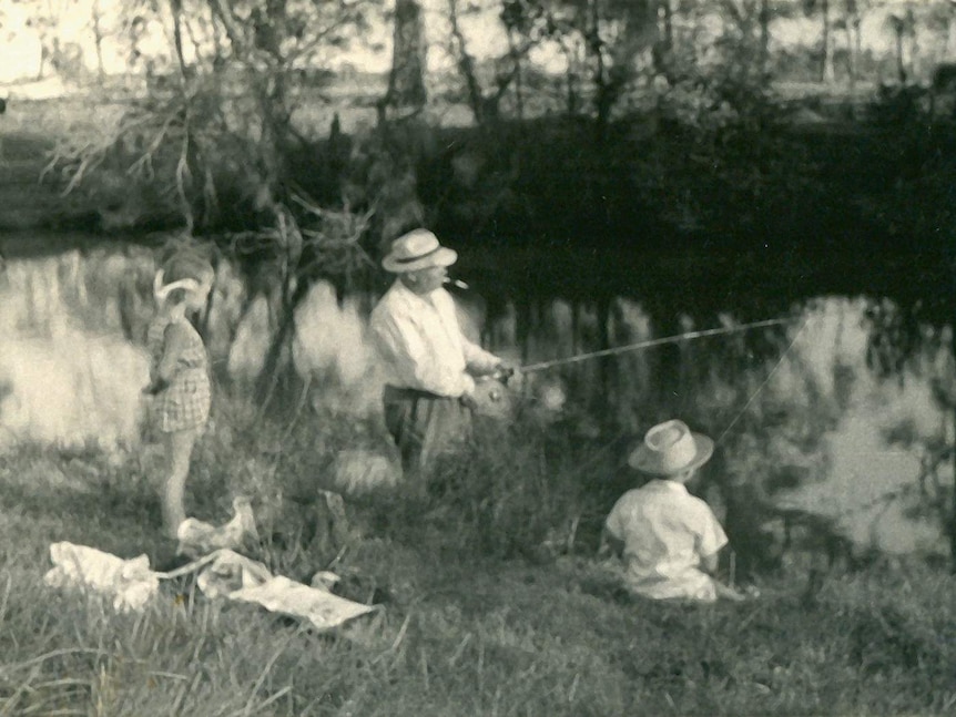 A blurry black and white photo, two children watch a man standing on the banks of a river, fishing.