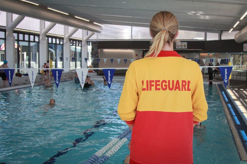 A lifeguard with long blonde hair keeps watch over an indoor swimming pool.