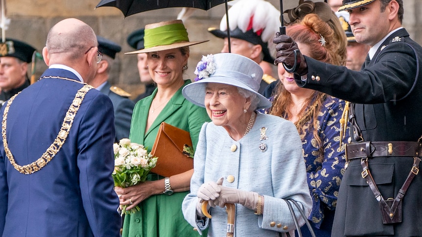 Queen Elizabeth smiles at an official holding a cane and dressed in light blue