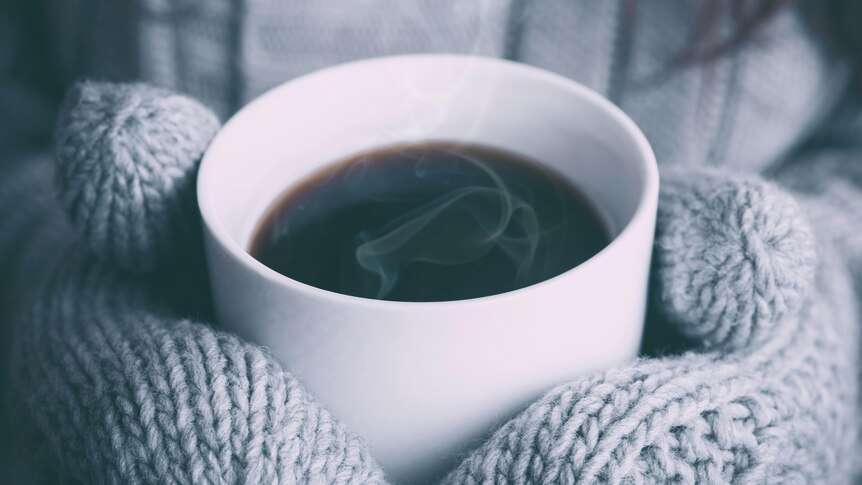 Two hands in heavy wool mittens holding a steaming cup of black coffee