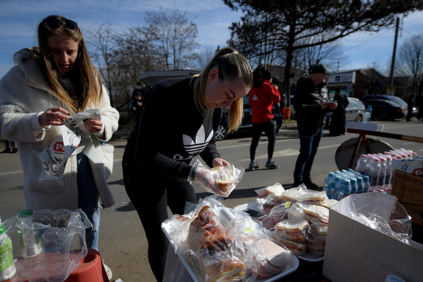 Two young females make sandwiches and wrap them individually and stack them along a trestle table.