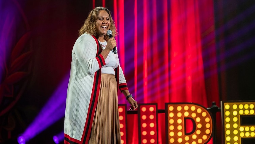 Elaine Crombie is performing on stage with a microphone in her hand. She wears a gold coloured skirt and a white cardigan. 