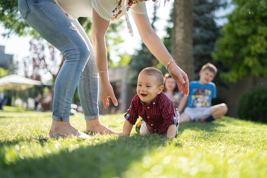 A woman bending over to pick up a crawling baby with older kids watching in the background