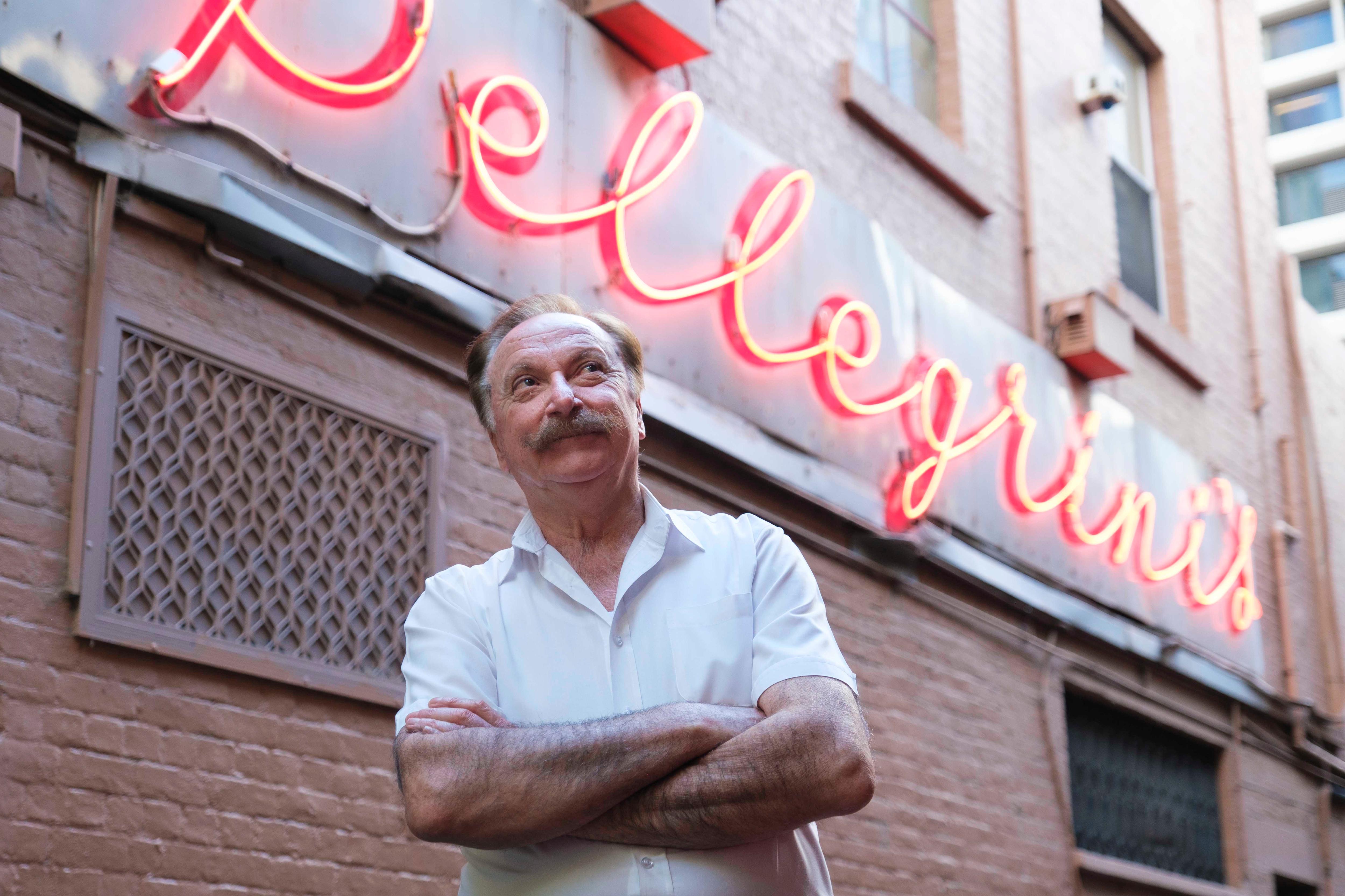 Rocco standing in front of a neon sign reading 'Pellegrini's' with his arms folded.