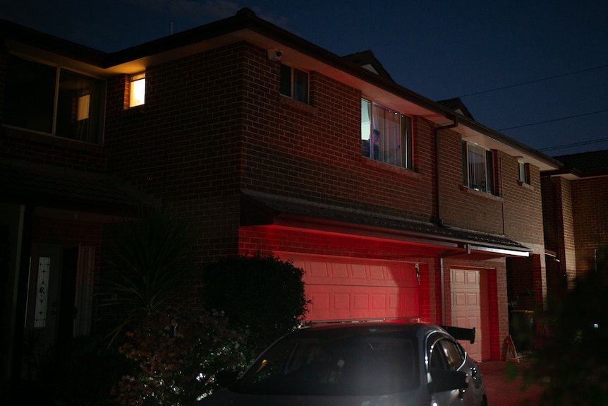 A young man looks out a window as a car drives away in the dark.