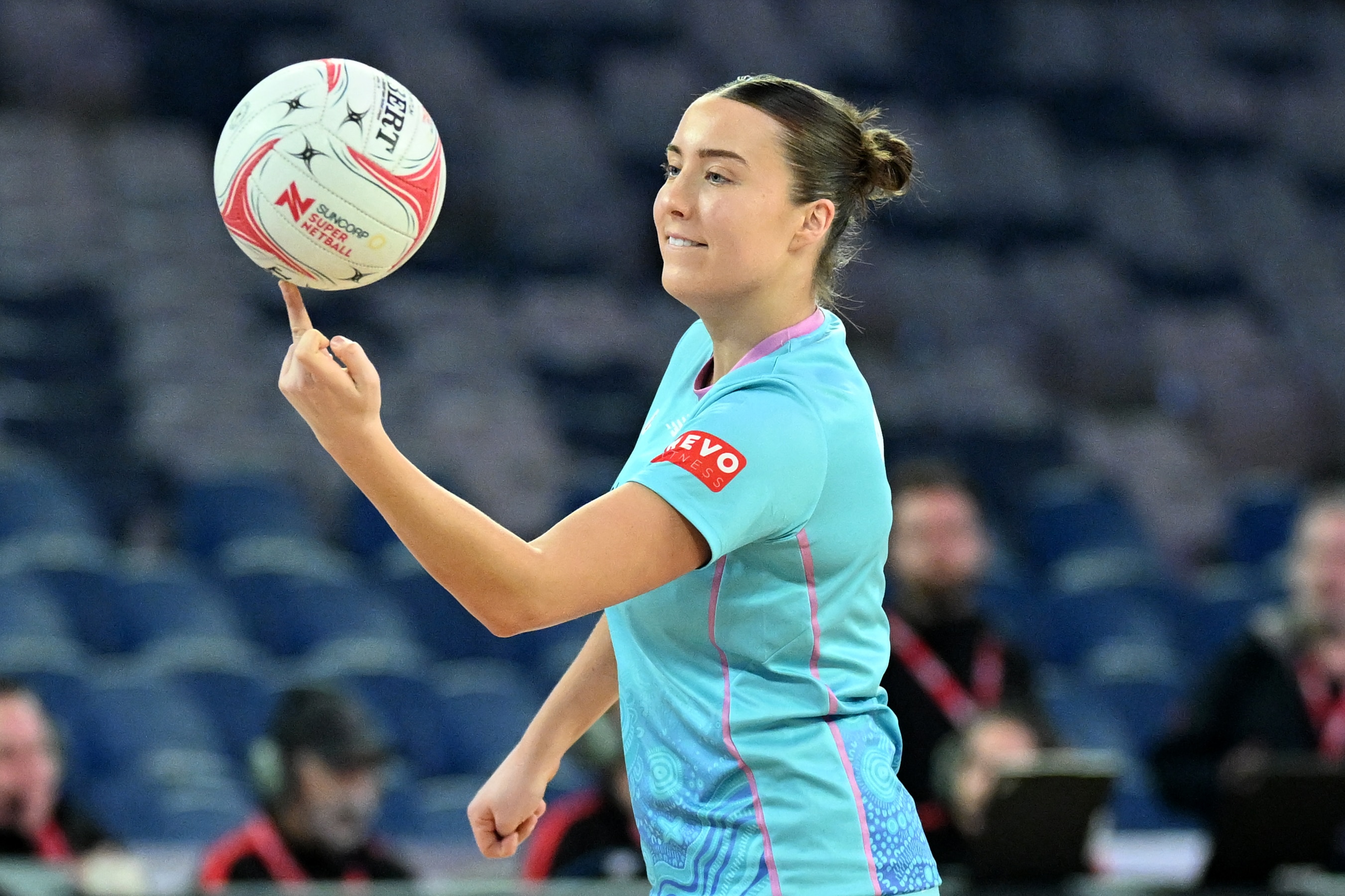 Wearing aqua, an athlete holds a netball up with her middle finger