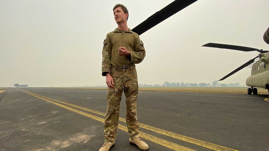 A soldier stands on a runway in uniform with a Chinook helicopter visible in the background.