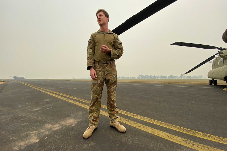 A soldier stands on a runway in uniform with a Chinook helicopter visible in the background.