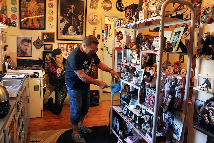 A man in a black Elvis Presley shirt dusting off a shelf of Elvis memorabilia.