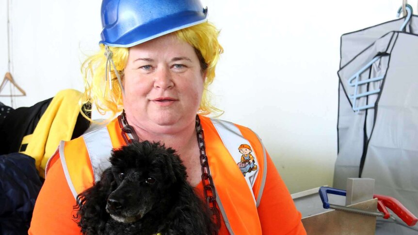 A lady dressed in hiviz and a hard hat holds a black poodle.