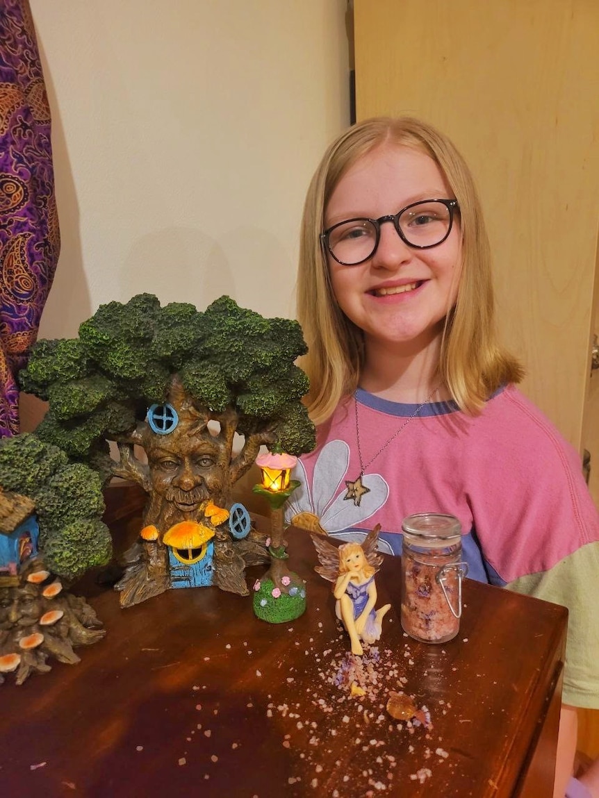 A young girl with glasses and a pink shirt standing behind a table with a fairy statue and fairy dust on the bench