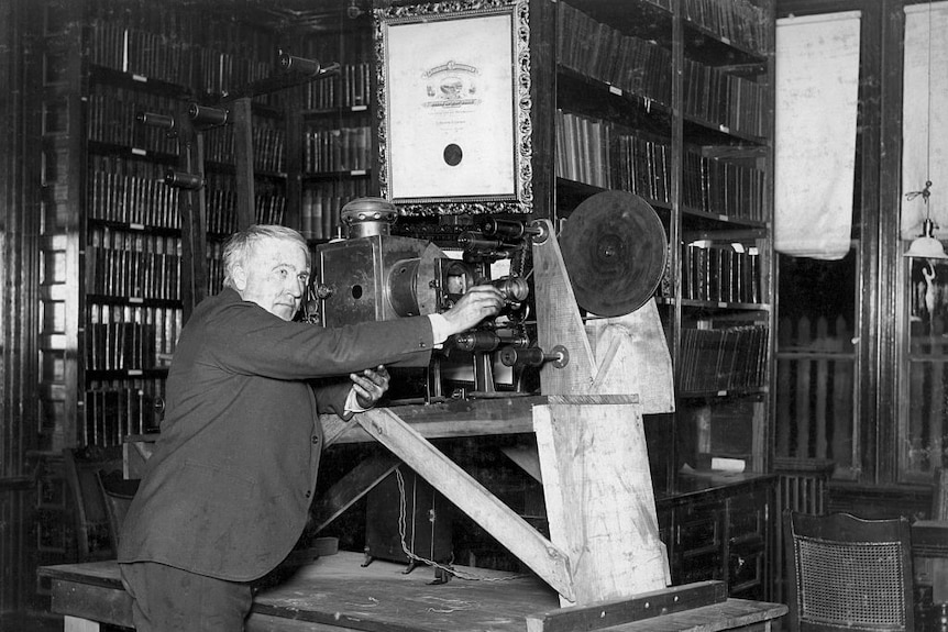 Black and white photograph of Thomas Edison in a suit looking through a projector machine