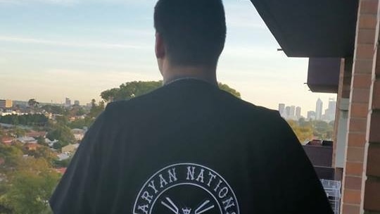 The back of a man wearing a black Aryan Nations t-shirt while standing on a balcony overlooking the Perth skyline.