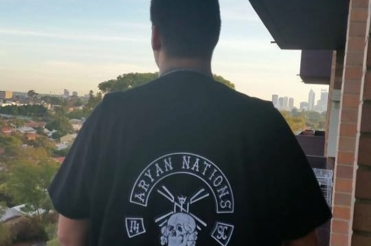 The back of a man wearing a black Aryan Nations t-shirt while standing on a balcony overlooking the Perth skyline.