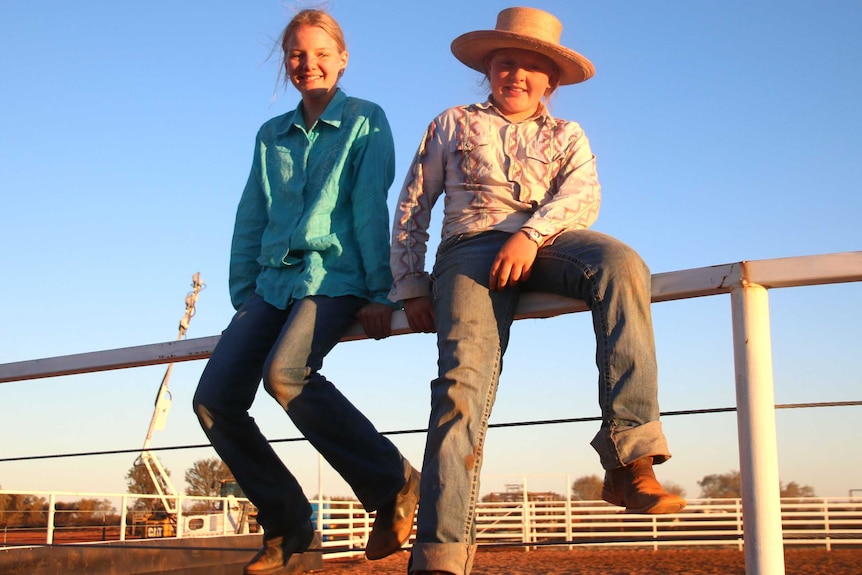 Two young girls sit on top of a rail of a rodeo arena.