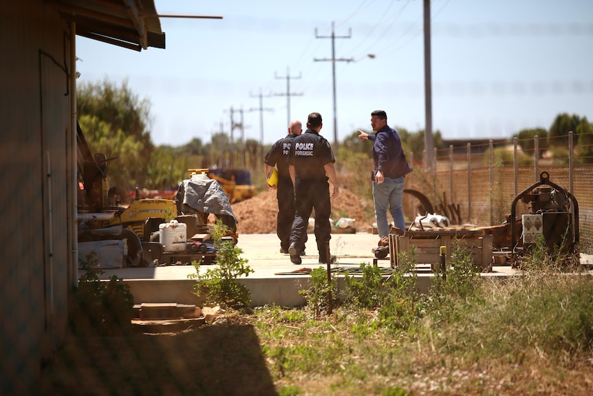 Two officers in forensic jumpsuits talk to a man in a workyard